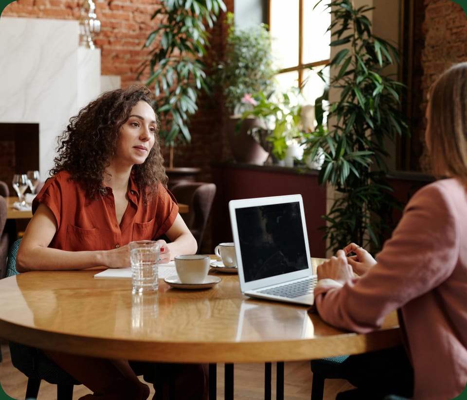 Two women talking across a café table, one listening with a laptop open